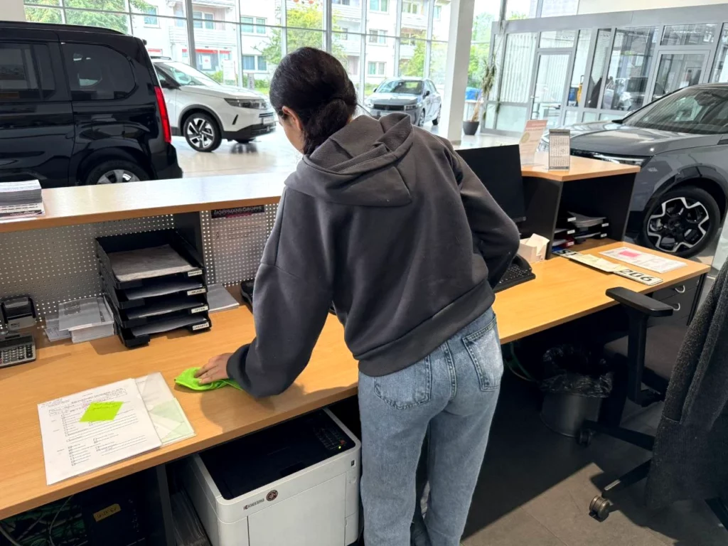 Eine Frau reinigt den Schreibtisch in einem Autohaus-Büro, im Hintergrund stehen mehrere Neuwagen.